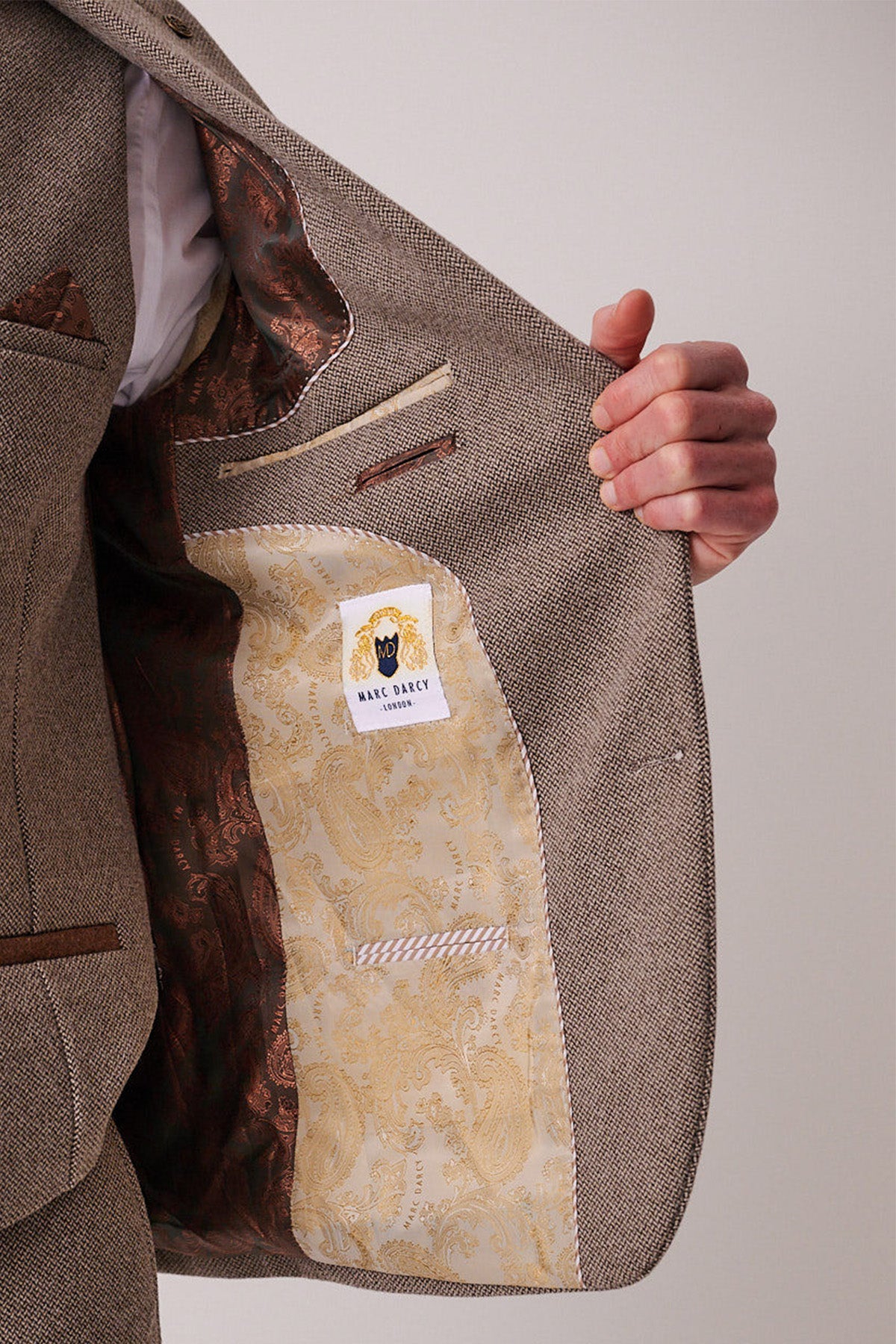 A person holds open the front of a marcdarcy HAYDEN Tan Crossweave Three Piece Suit blazer, showing its ornate golden paisley lining and a label featuring the marcdarcy logo of a man in a hat.