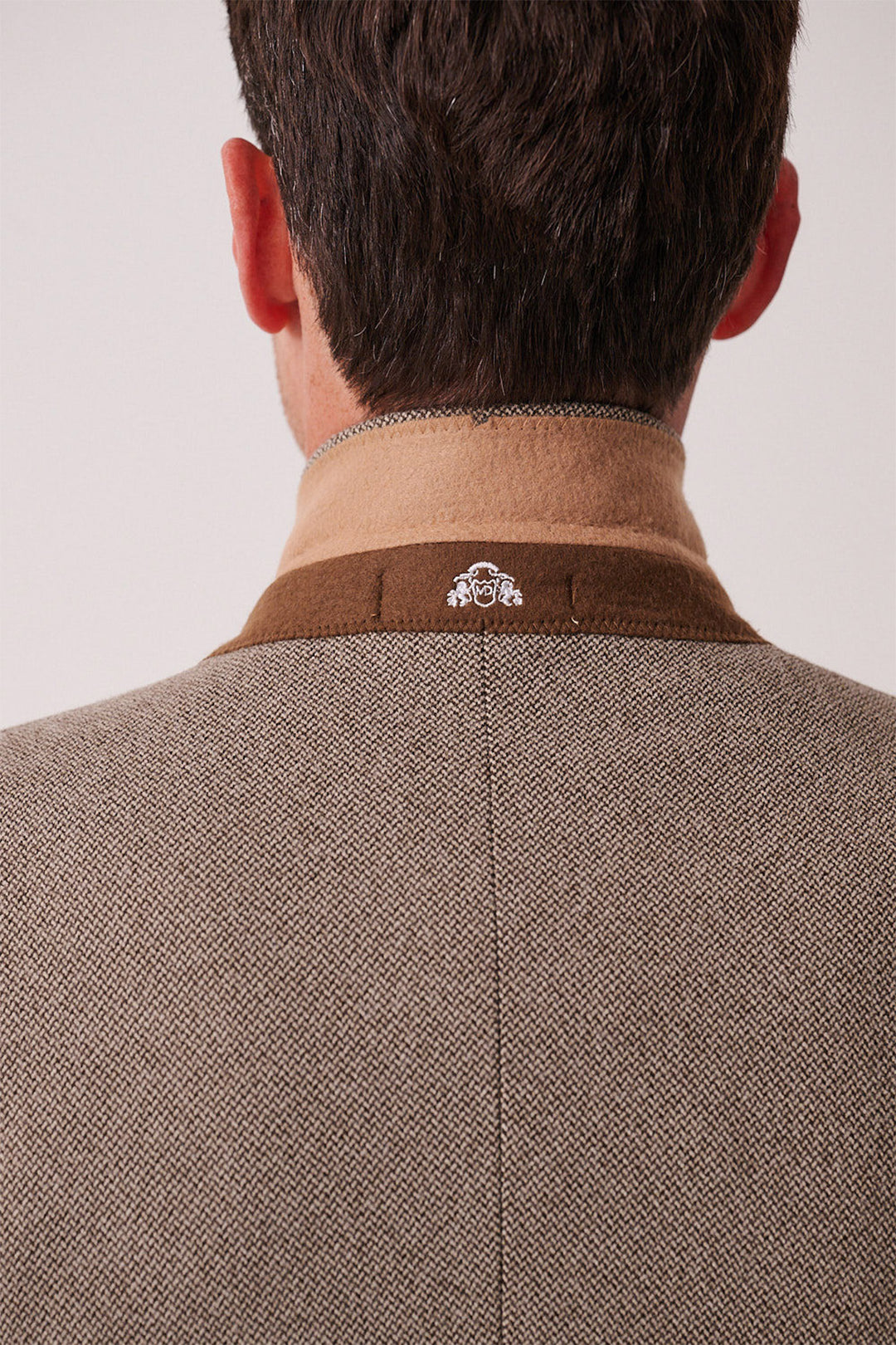 A close-up of the back of a man's head and shoulders showcases the marcdarcy HAYDEN - Tan Crossweave Three Piece Suit, featuring a small white embroidered logo on the collar against a softly lit, plain background.