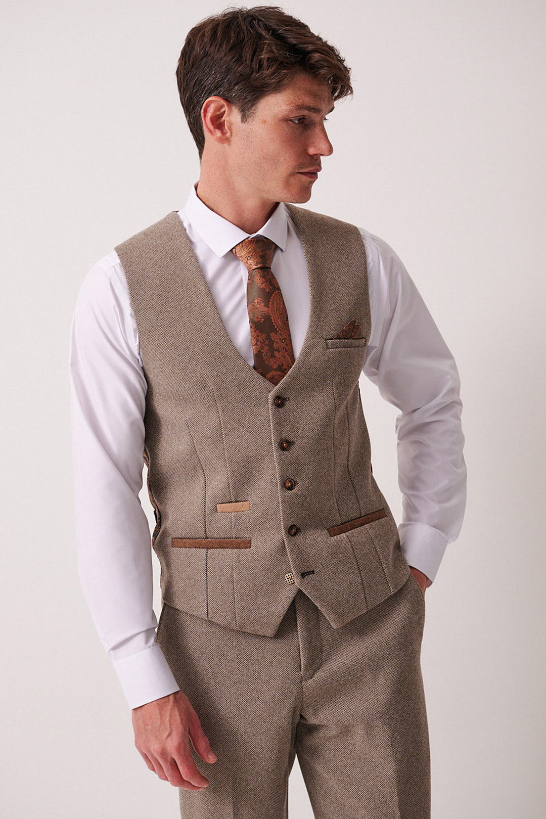A man in the marcdarcy HAYDEN Tan Crossweave Three Piece Suit with a matching vest and trousers, a white shirt, and a brown patterned tie stands against a plain light background, looking left with one hand in his pocket.