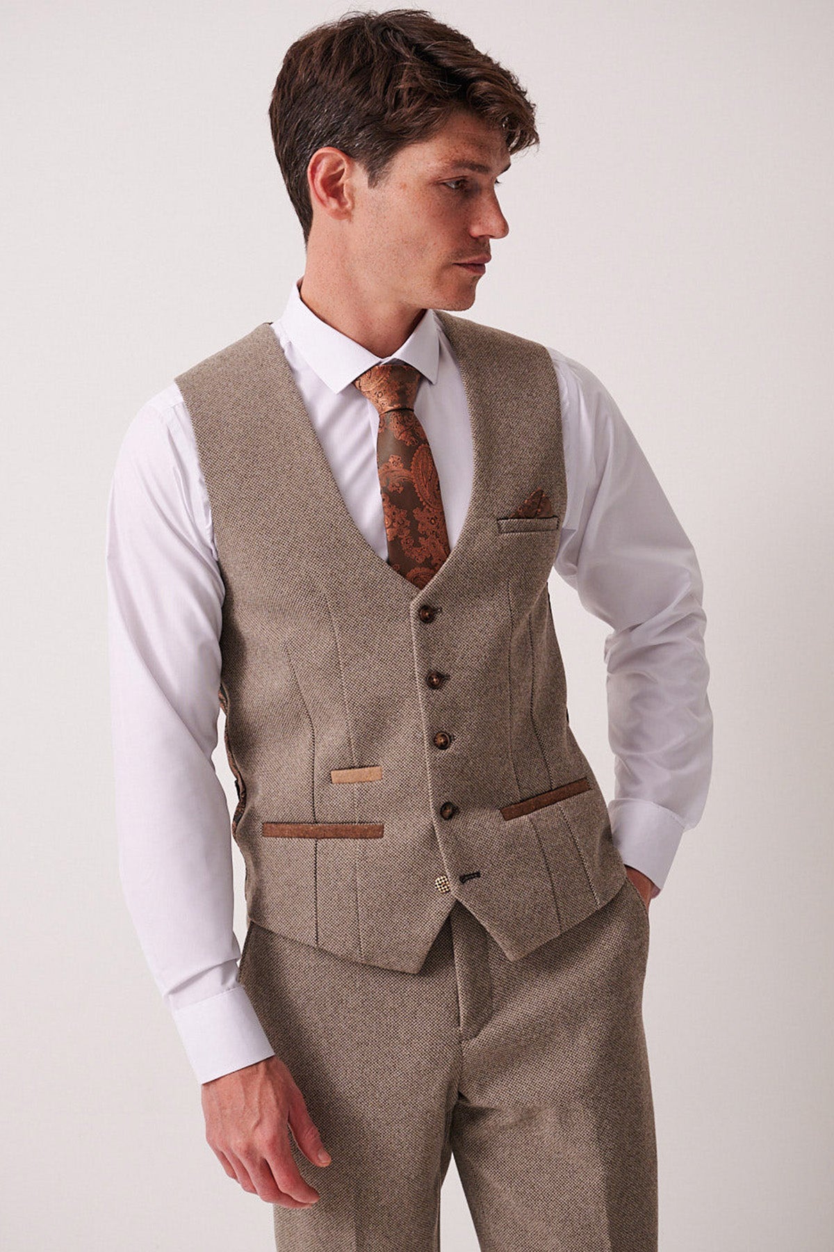 A man in the marcdarcy HAYDEN Tan Crossweave Three Piece Suit with a matching vest and trousers, a white shirt, and a brown patterned tie stands against a plain light background, looking left with one hand in his pocket.