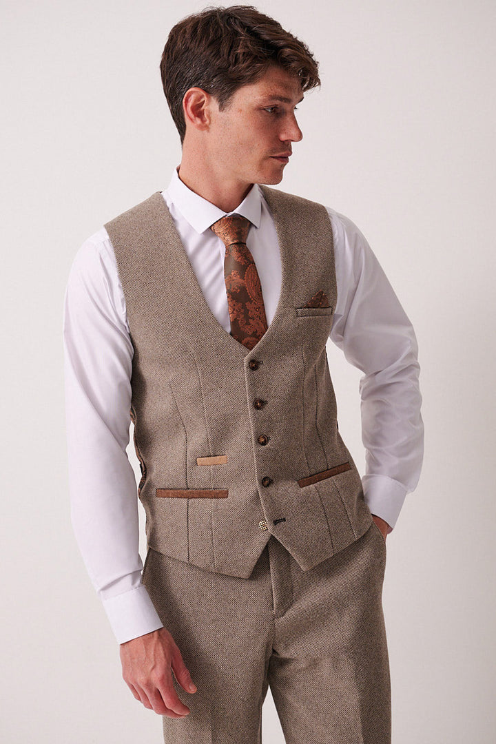 A man in the marcdarcy HAYDEN Tan Crossweave Three Piece Suit with a matching vest and trousers, a white shirt, and a brown patterned tie stands against a plain light background, looking left with one hand in his pocket.