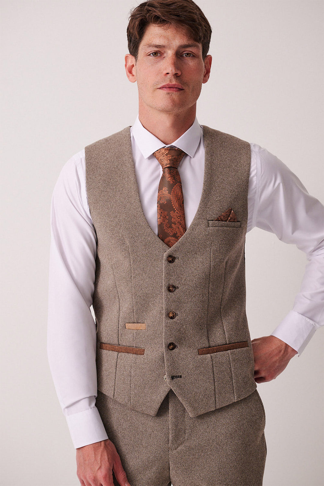 A man poses confidently against a light background wearing the marcdarcy HAYDEN Tan Crossweave Three Piece Suit, styled with a white dress shirt and patterned brown tie, one arm bent.