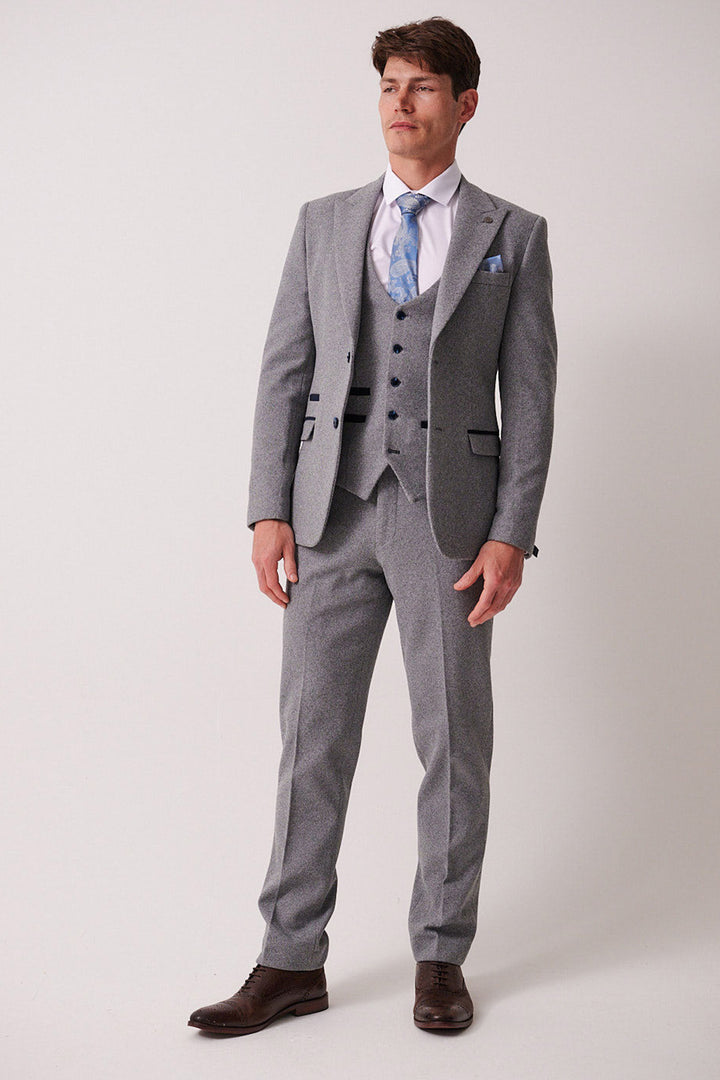 A man stands against a plain background wearing the marcdarcy HAYDEN Silver Blue Crossweave Three Piece Suit, styled with a white shirt, light blue tie, matching pocket square, and brown dress shoes.