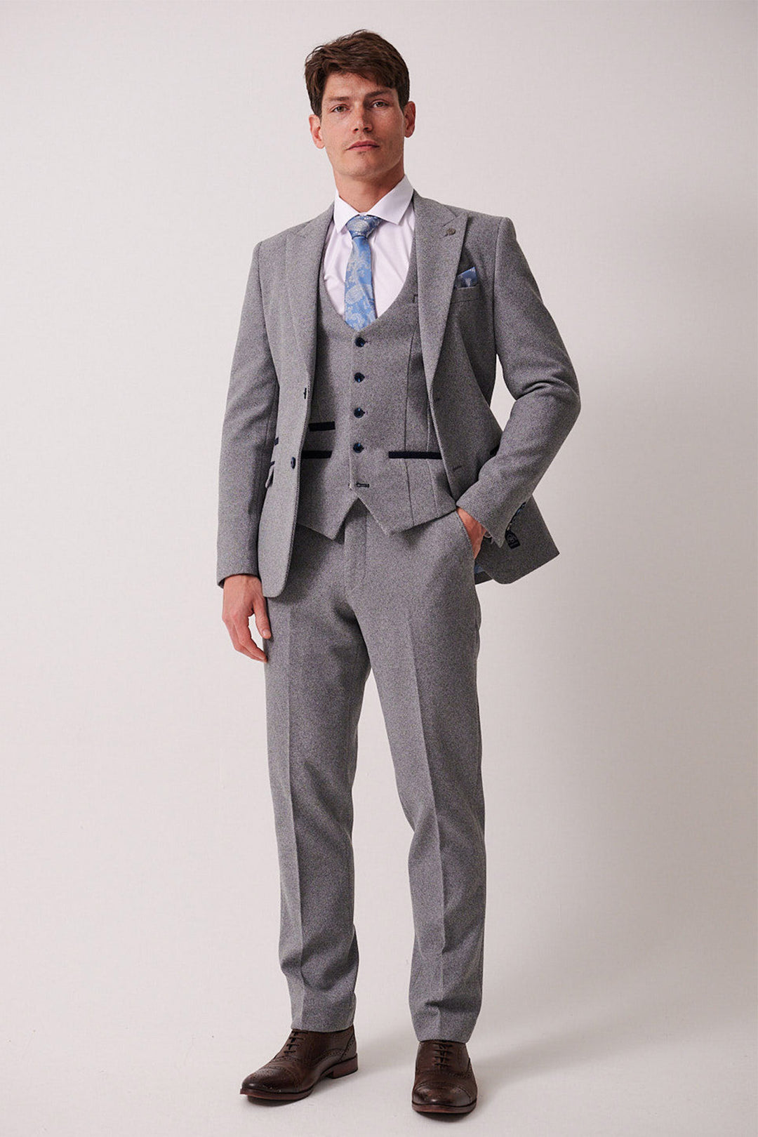 A man wears the marcdarcy HAYDEN - Silver Blue Crossweave Three Piece Suit with a white shirt, light blue patterned tie, and brown dress shoes against a plain background.