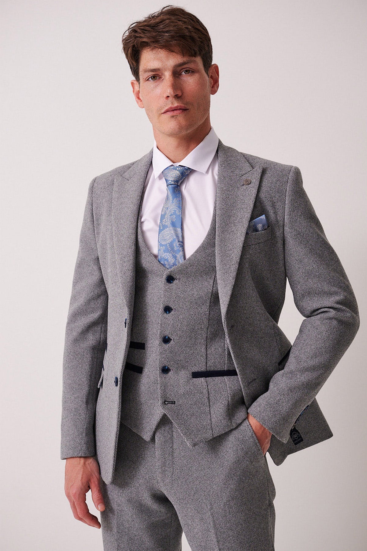 A man stands against a plain background wearing the marcdarcy HAYDEN Silver Blue Crossweave Blazer, a white dress shirt, and a light blue patterned tie. His right hand is in his pocket and he has a neutral expression.