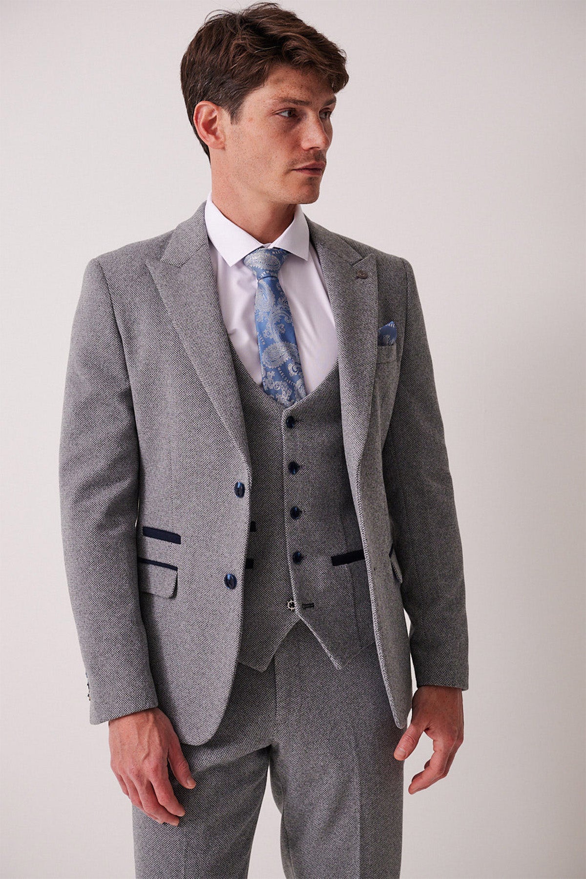 A man in the marcdarcy HAYDEN Silver Blue Crossweave Three Piece Suit, paired with a white shirt and blue patterned tie, stands against a plain light background, looking to his left.