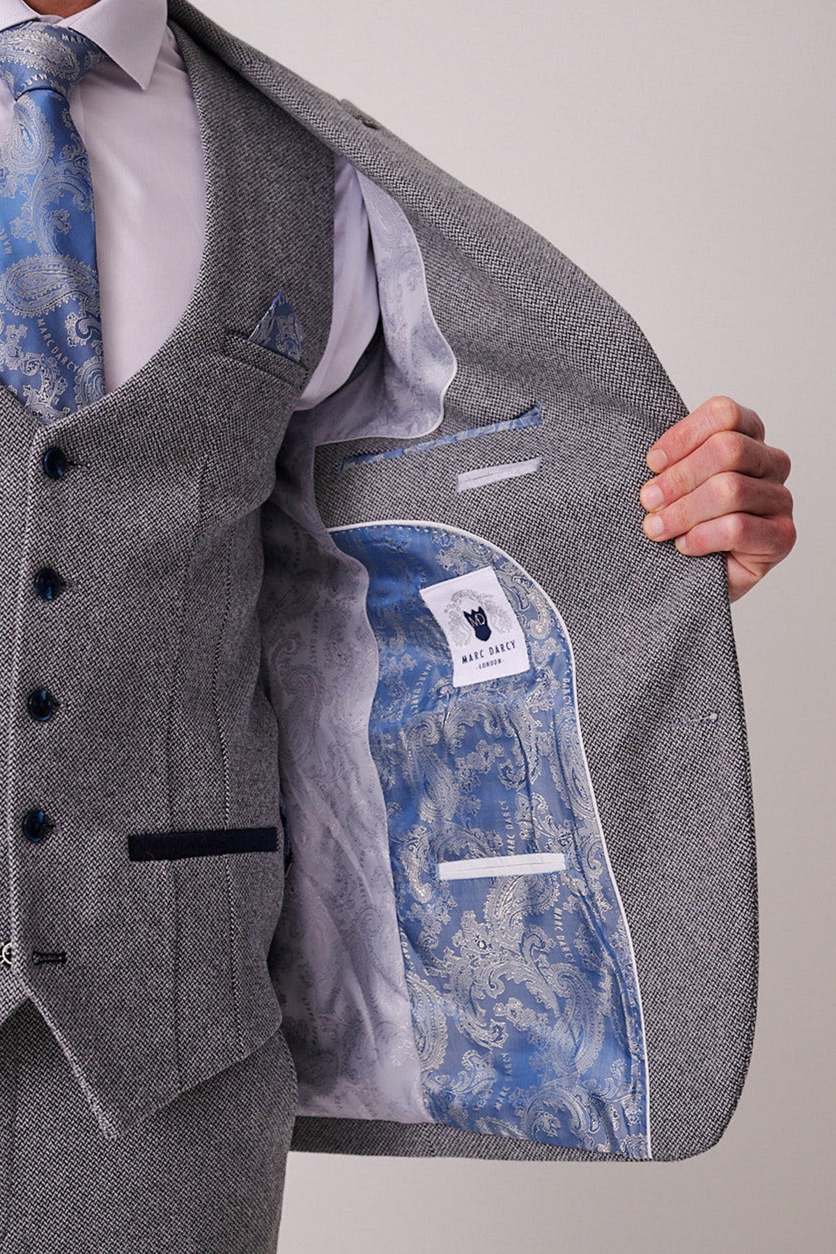 A person in a textured marcdarcy HAYDEN - Silver Blue Crossweave Blazer holds open their jacket to reveal a light blue patterned lining, matching waistcoat, blue paisley tie, and the label inside the men's blazer.