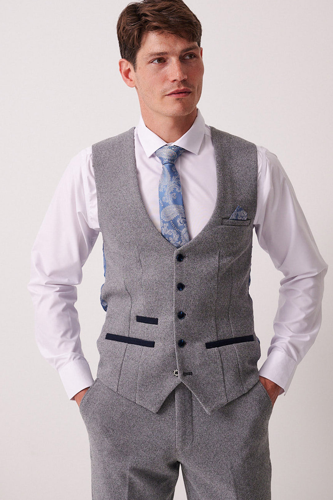 A man wears the Marc Darcy HAYDEN Silver Blue Crossweave Waistcoat with matching trousers, a white shirt, and a blue patterned tie. He has short brown hair, stands with hands in pockets, and gazes slightly to the side.