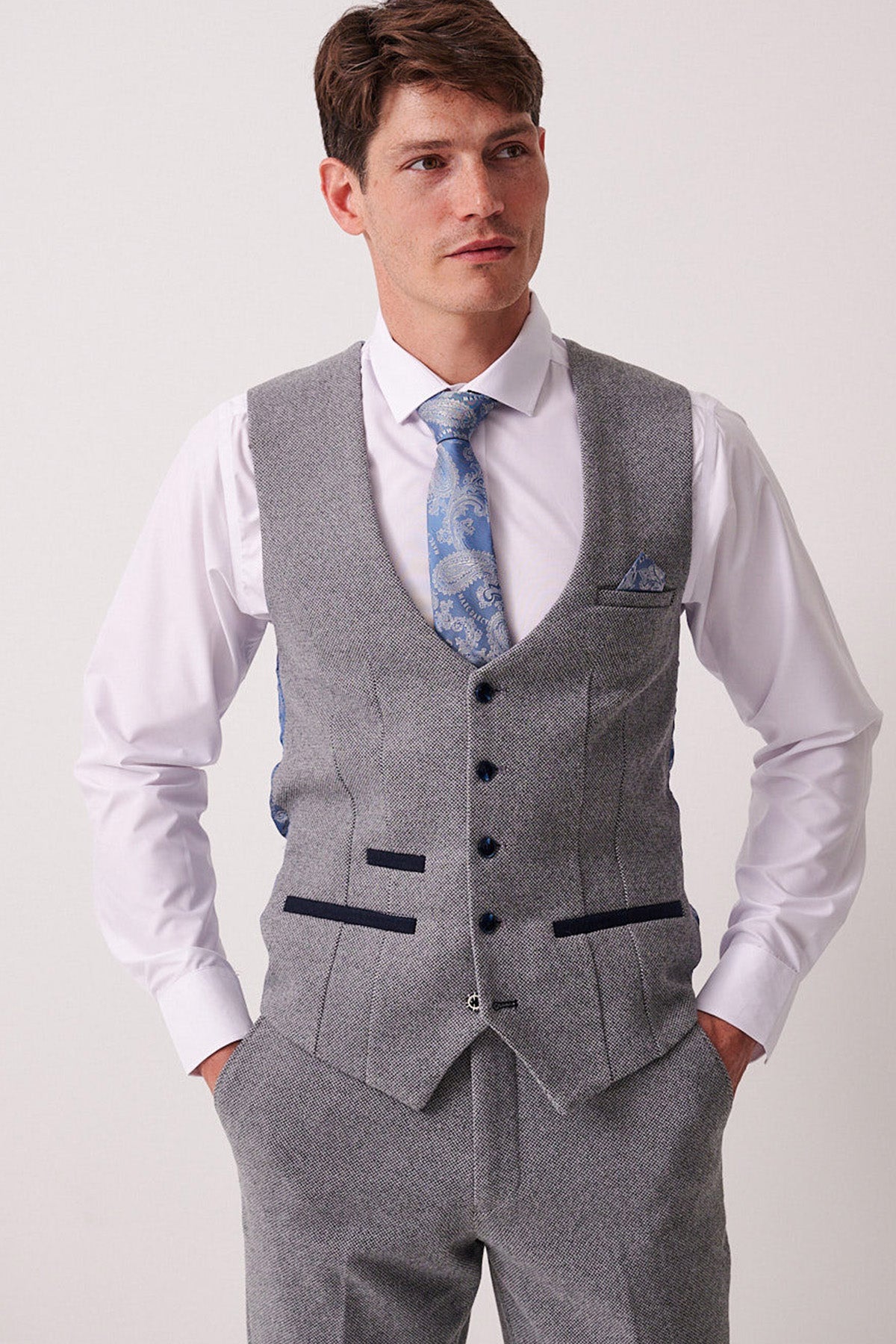 A man wears the Marc Darcy HAYDEN Silver Blue Crossweave Waistcoat with matching trousers, a white shirt, and a blue patterned tie. He has short brown hair, stands with hands in pockets, and gazes slightly to the side.