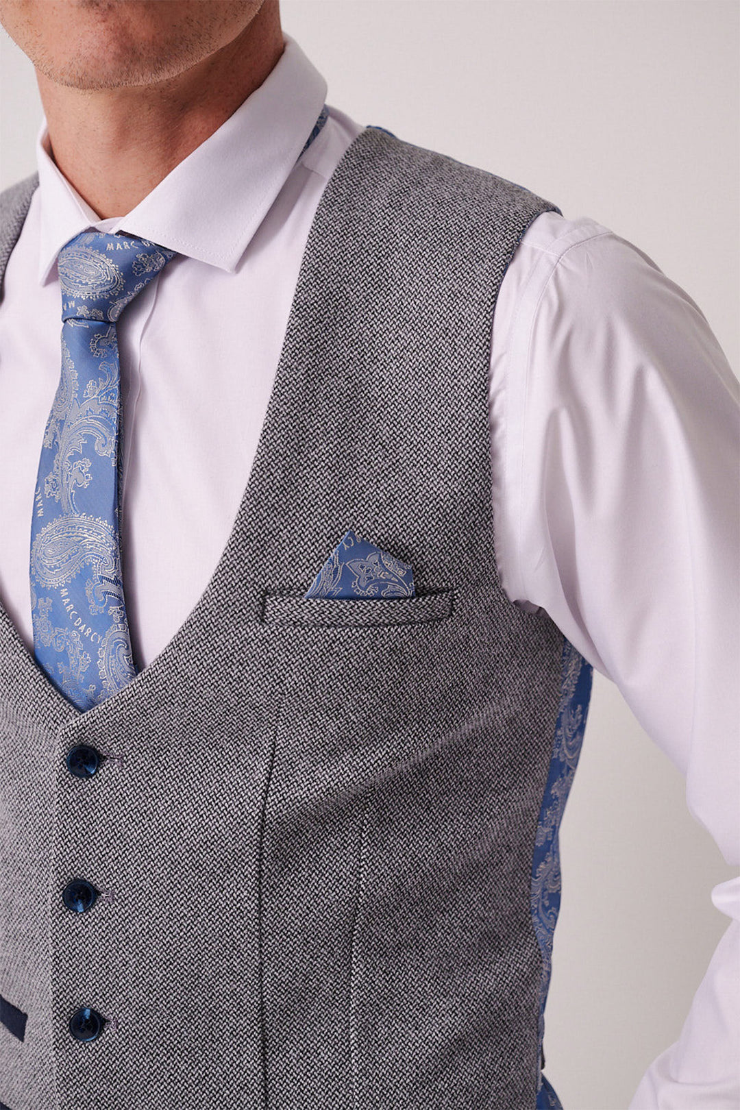 A man wears the marcdarcy HAYDEN Silver Blue Crossweave Three Piece Suit, featuring a light gray textured waistcoat, white shirt, and blue patterned tie with matching pocket square, styled slim fit against a plain background.