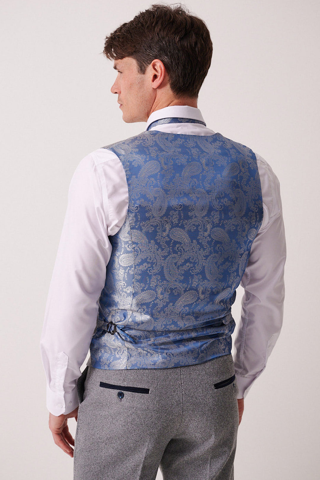 A man faces away, wearing the marcdarcy HAYDEN Silver Blue Crossweave Three Piece Suit: a slim fit waistcoat over a white shirt and light gray trousers with dark pocket trim.