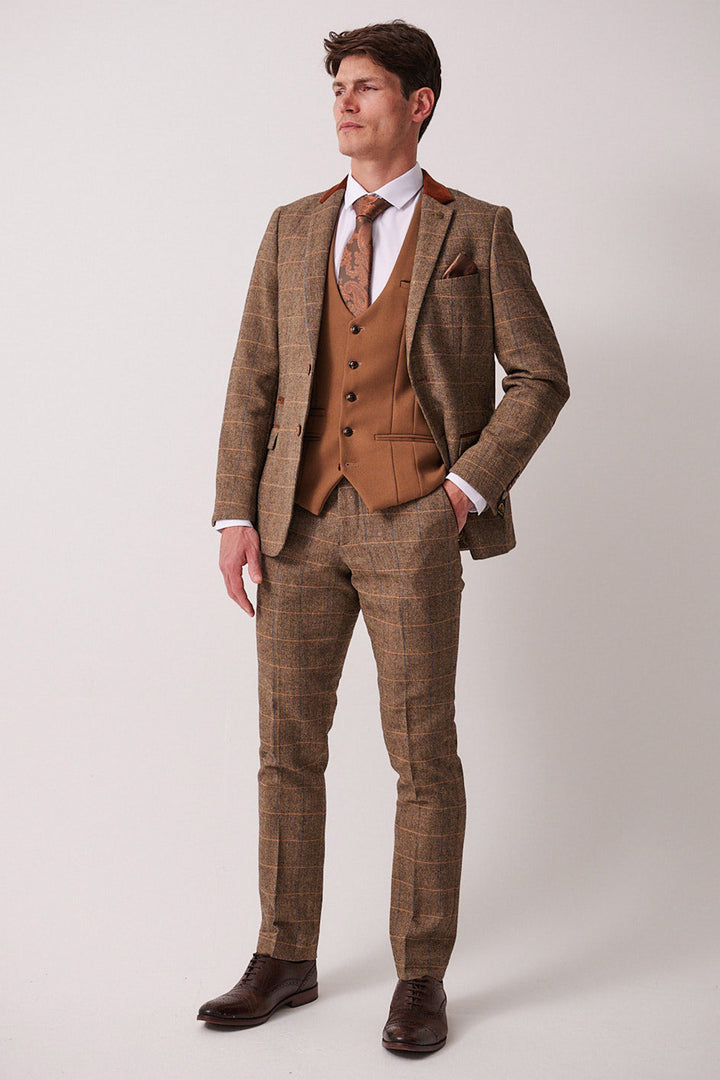 A man with short brown hair stands against a plain background wearing the marcdarcy DX7 - Tan Tweed Check Suit With Kelvin Oak Waistcoat, styled with a white shirt, brown tie, and dark brown shoes—ideal for a country wedding. One hand is in his pocket.