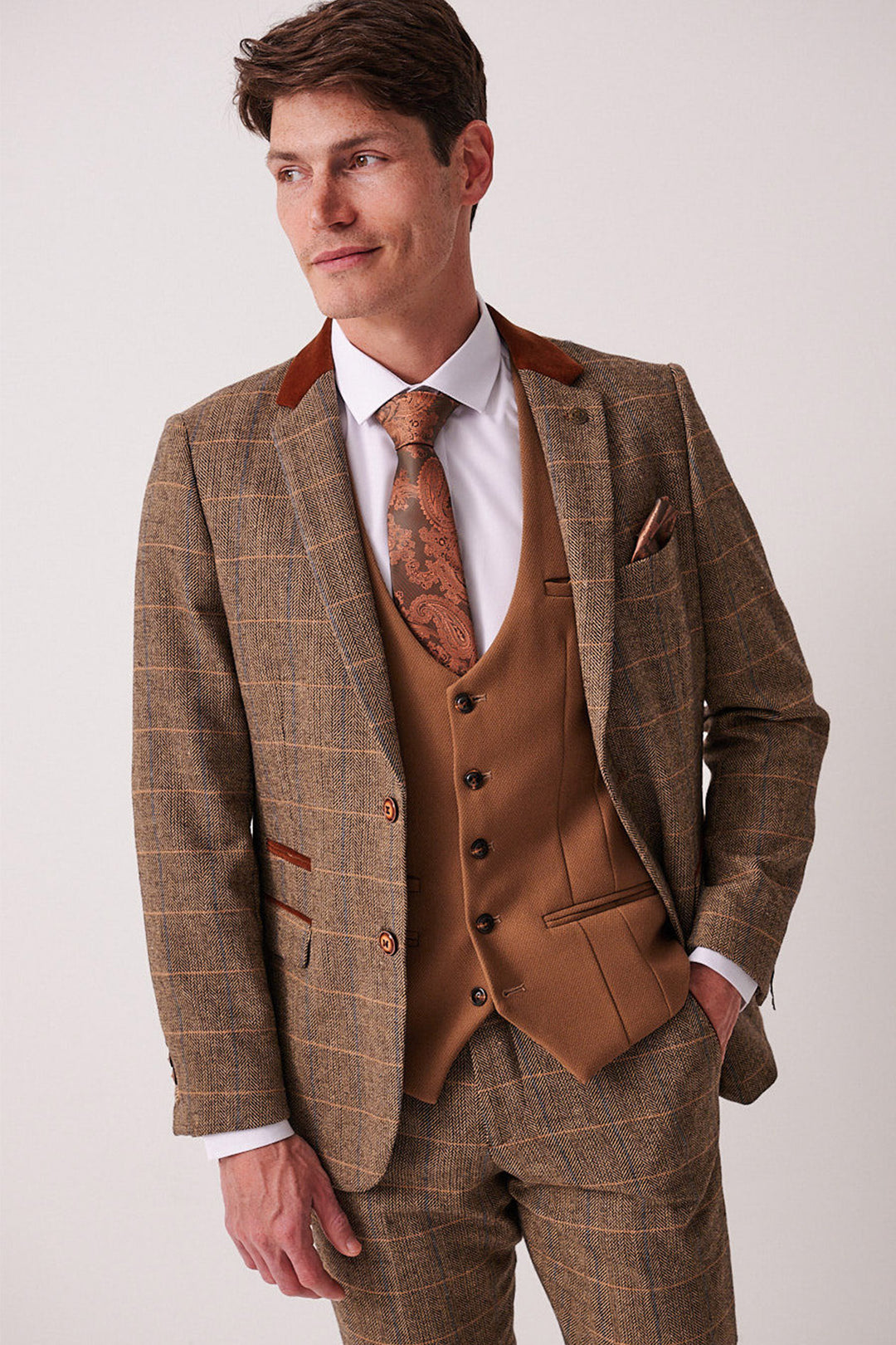 A man in the marcdarcy DX7 Tan Tweed Check Suit with Kelvin Oak waistcoat, white shirt, and brown patterned tie stands with one hand in his pocket against a plain background. Perfect for a country wedding.