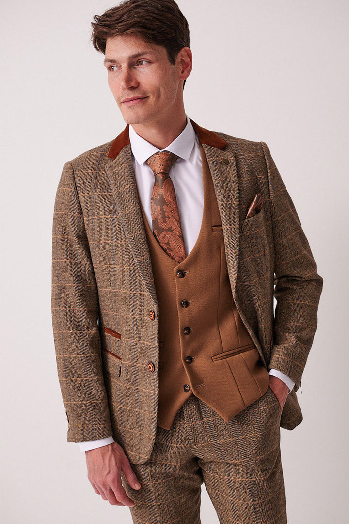 A man in the marcdarcy DX7 Tan Tweed Check Suit with Kelvin Oak waistcoat, white shirt, and brown patterned tie stands with one hand in his pocket against a plain background. Perfect for a country wedding.