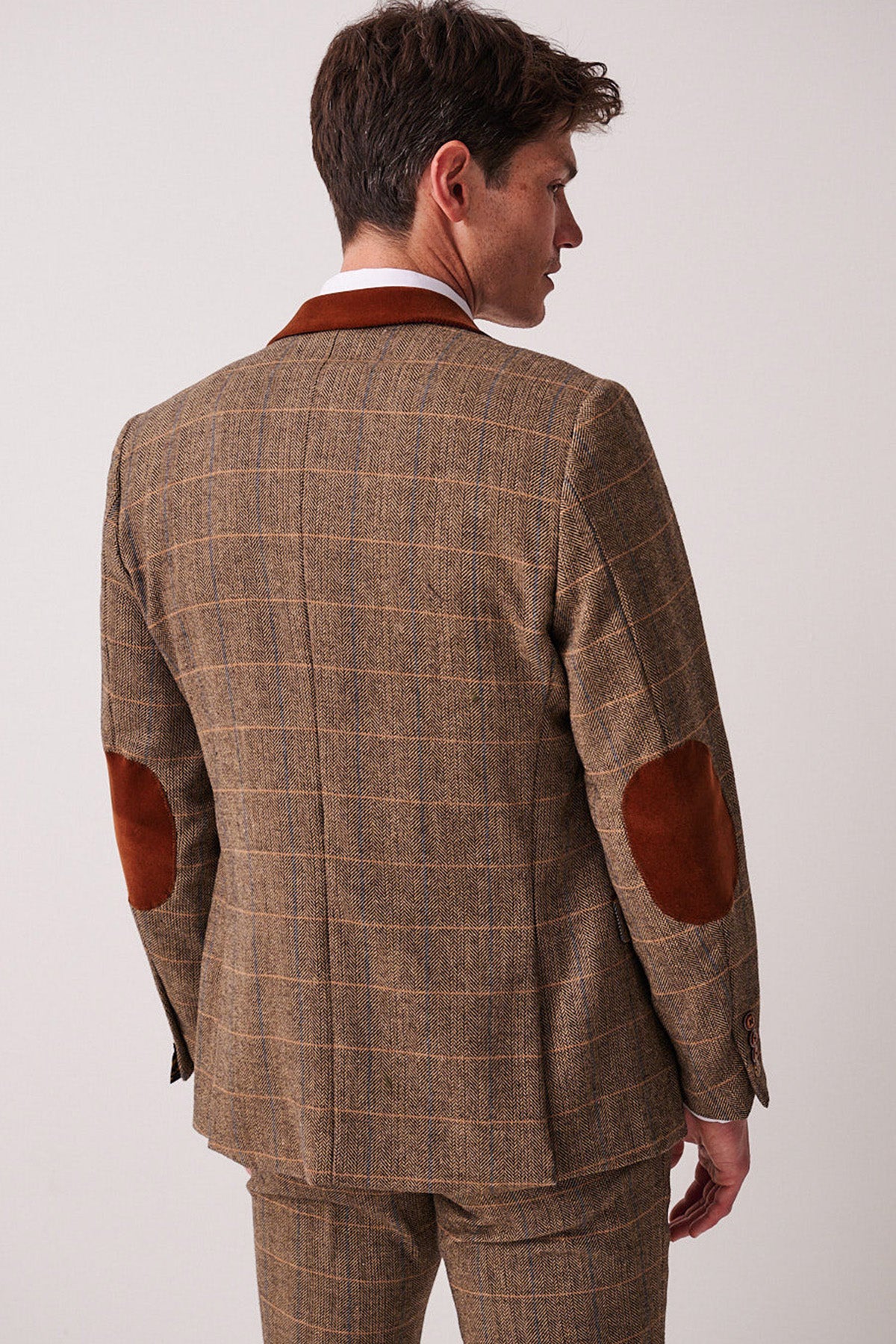Dressed in the marcdarcy DX7 Tan Tweed Check Suit with Kelvin Oak Waistcoat, a man stands facing away, glancing over his shoulder. The light background highlights this timeless style—ideal for a country wedding.