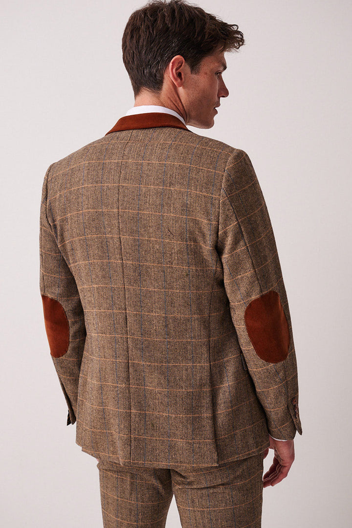 Dressed in the marcdarcy DX7 Tan Tweed Check Suit with Kelvin Oak Waistcoat, a man stands facing away, glancing over his shoulder. The light background highlights this timeless style—ideal for a country wedding.