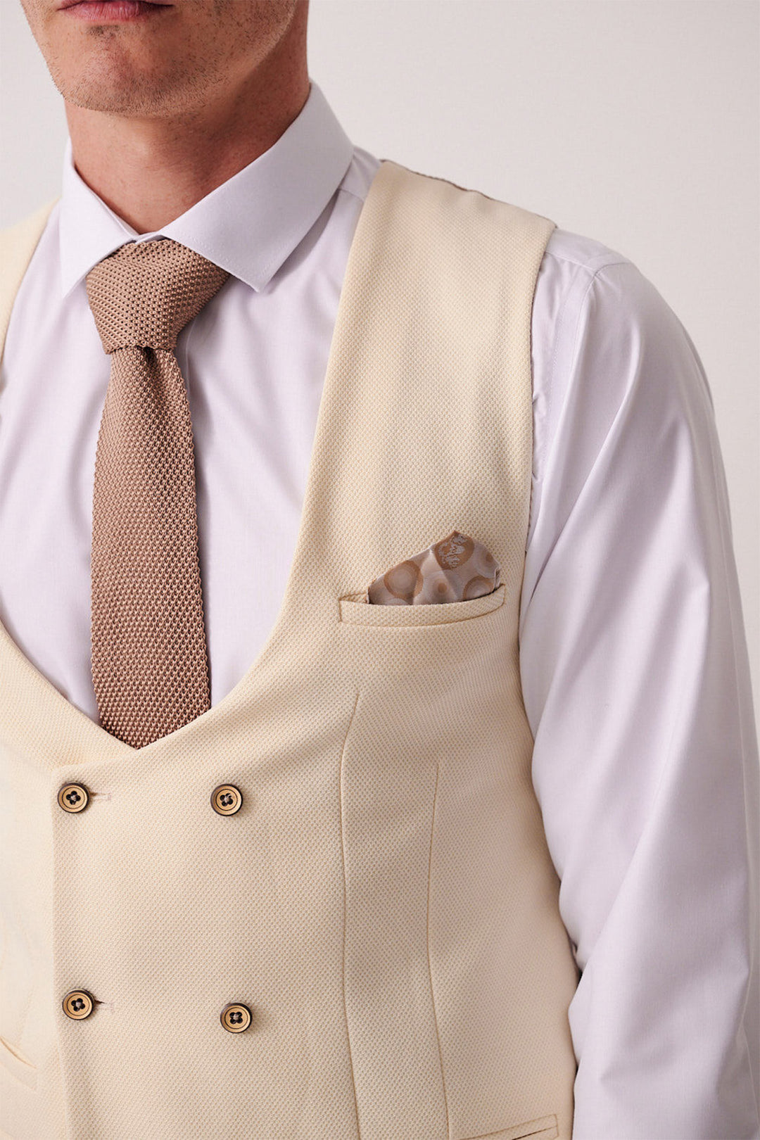 A man wears a marcdarcy DX7 Tan Tweed Check Suit with a Kelvin Cream Waistcoat, white shirt, textured brown tie, and circular pattern pocket square for a vintage-inspired look. Only his upper torso and part of his face are visible.