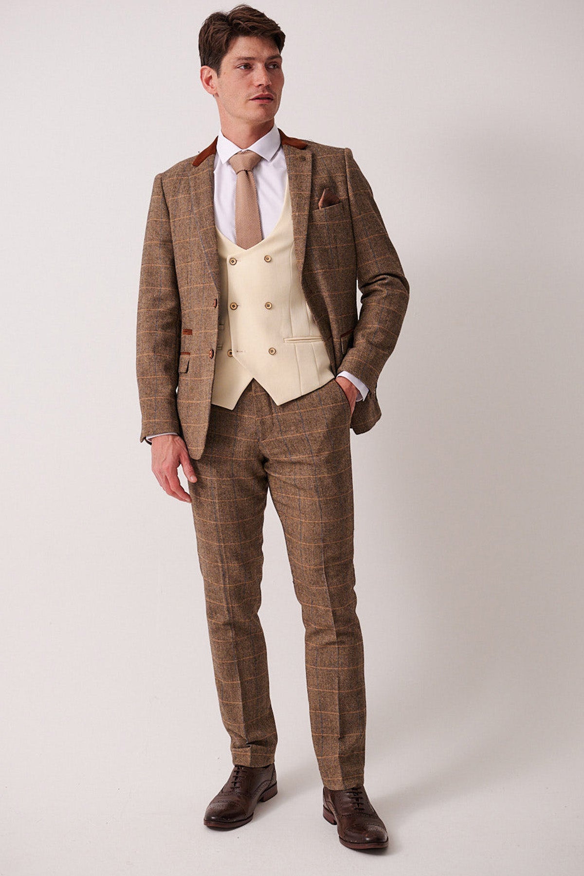 A man stands against a plain background wearing the marcdarcy DX7 - Tan Tweed Check Suit with Kelvin Cream Waistcoat, paired with a white shirt, beige tie, and dark brown shoes. He looks slightly to the side with one hand in his pocket.