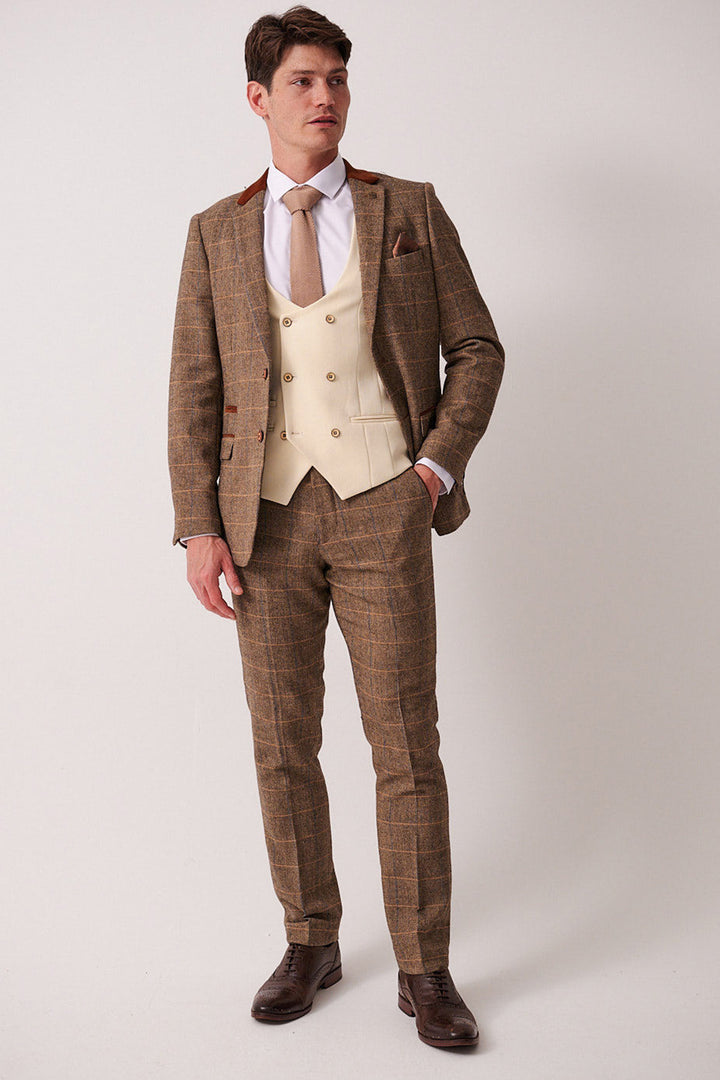 A man stands against a plain background wearing the marcdarcy DX7 - Tan Tweed Check Suit with Kelvin Cream Waistcoat, paired with a white shirt, beige tie, and dark brown shoes. He looks slightly to the side with one hand in his pocket.