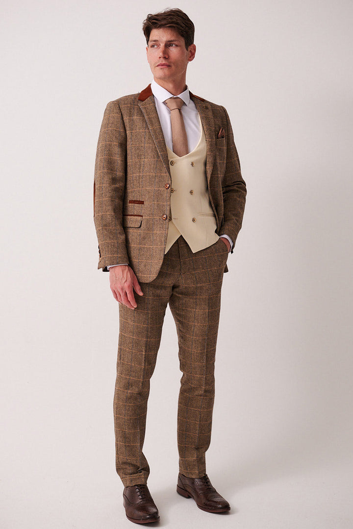 A man poses in the marcdarcy DX7 Tan Tweed Check Suit with Kelvin Cream Waistcoat, styled with a tan tie, herringbone slim fit blazer, brown leather shoes, and a tan pocket square.