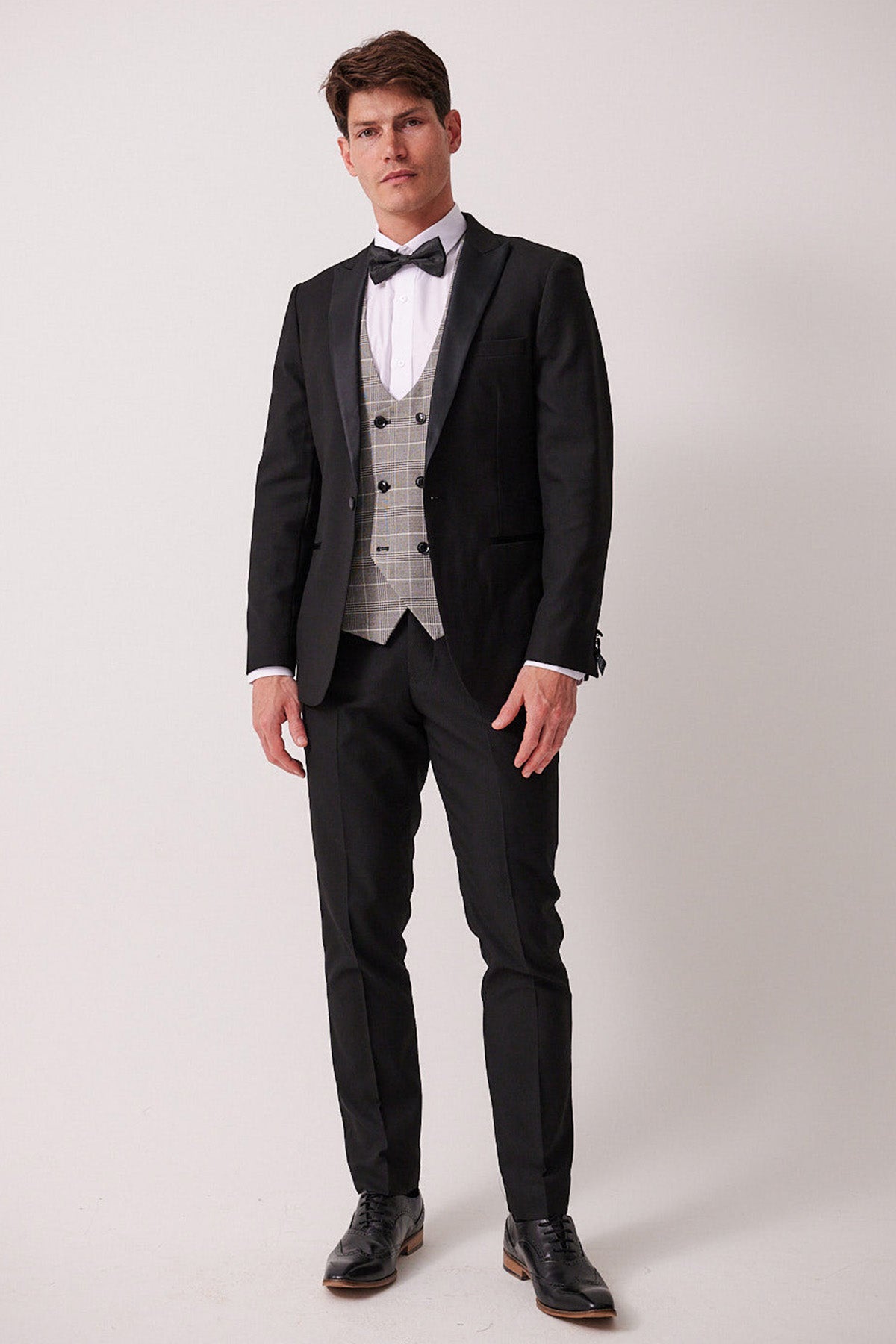 A man wears the marcdarcy DALTON Black Tux Lapel Suit with a Ross Waistcoat, white shirt, black bow tie, black dress pants, and polished black wingtip shoes.