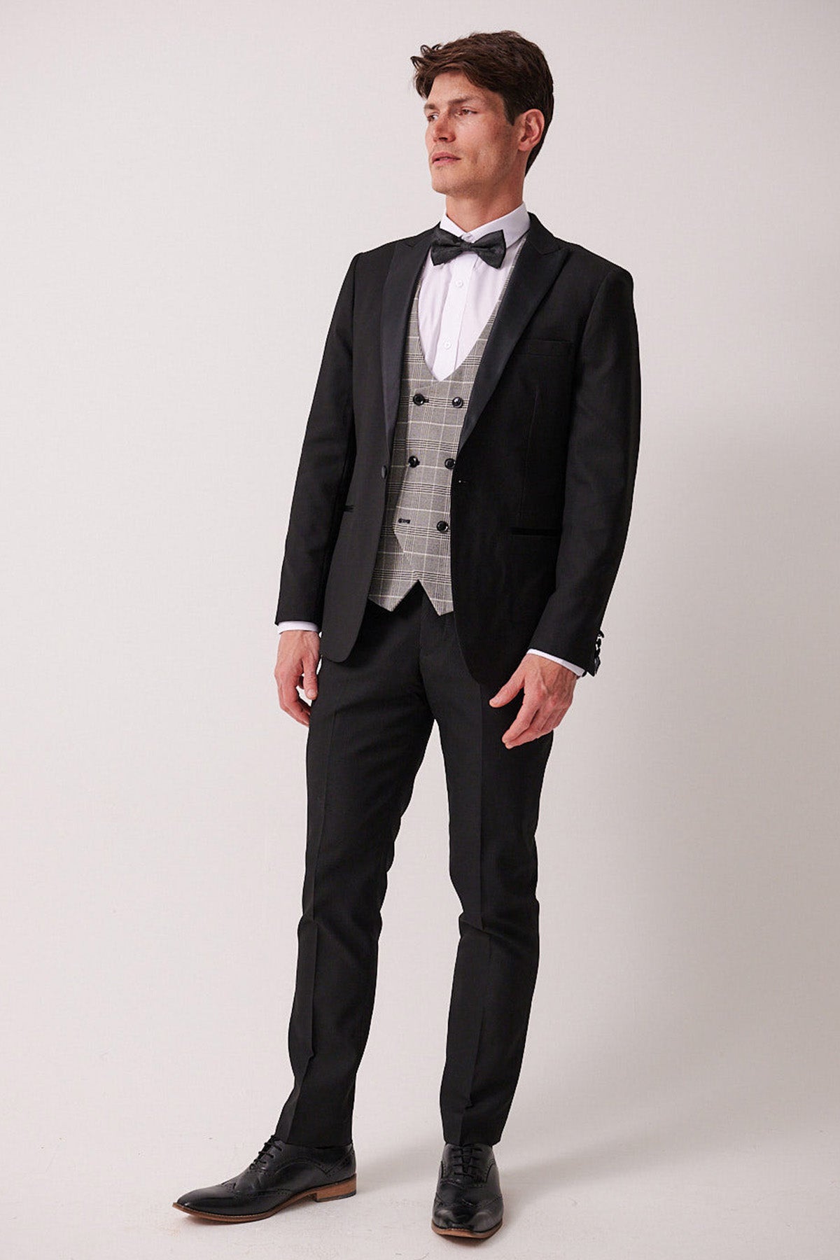 A man in the marcdarcy DALTON Black Tux Lapel Suit with Ross Waistcoat stands against a plain light background, wearing a slim fit blazer, white shirt, black bow tie, and light gray plaid waistcoat, looking slightly to the side.