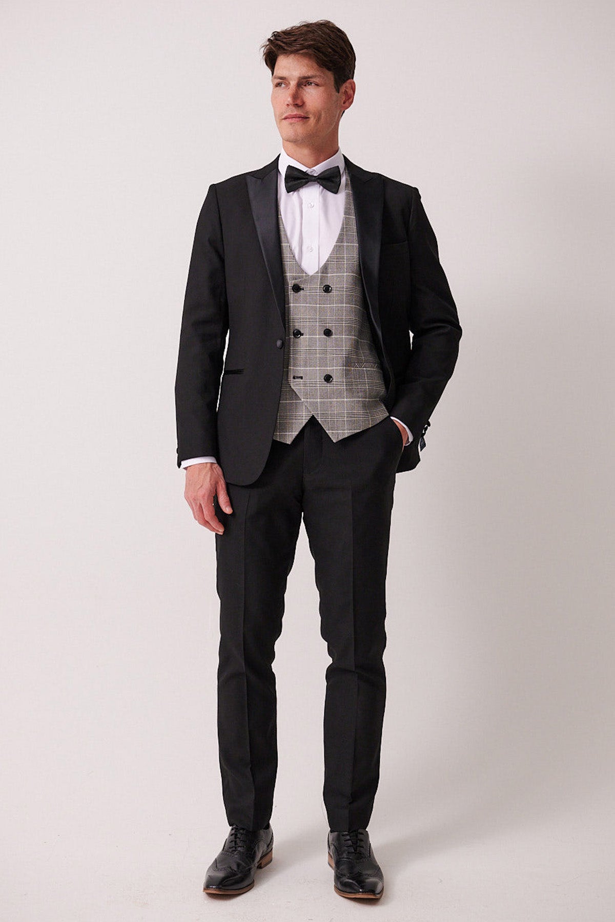 A man with short brown hair stands against a plain background wearing marcdarcy’s DALTON – Black Tux Lapel Suit with Ross Waistcoat: a slim fit blazer, black tie, white shirt, gray plaid vest, and black dress shoes.