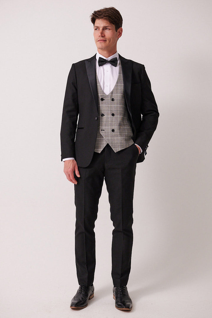 A man with short brown hair stands against a plain background wearing marcdarcy’s DALTON – Black Tux Lapel Suit with Ross Waistcoat: a slim fit blazer, black tie, white shirt, gray plaid vest, and black dress shoes.