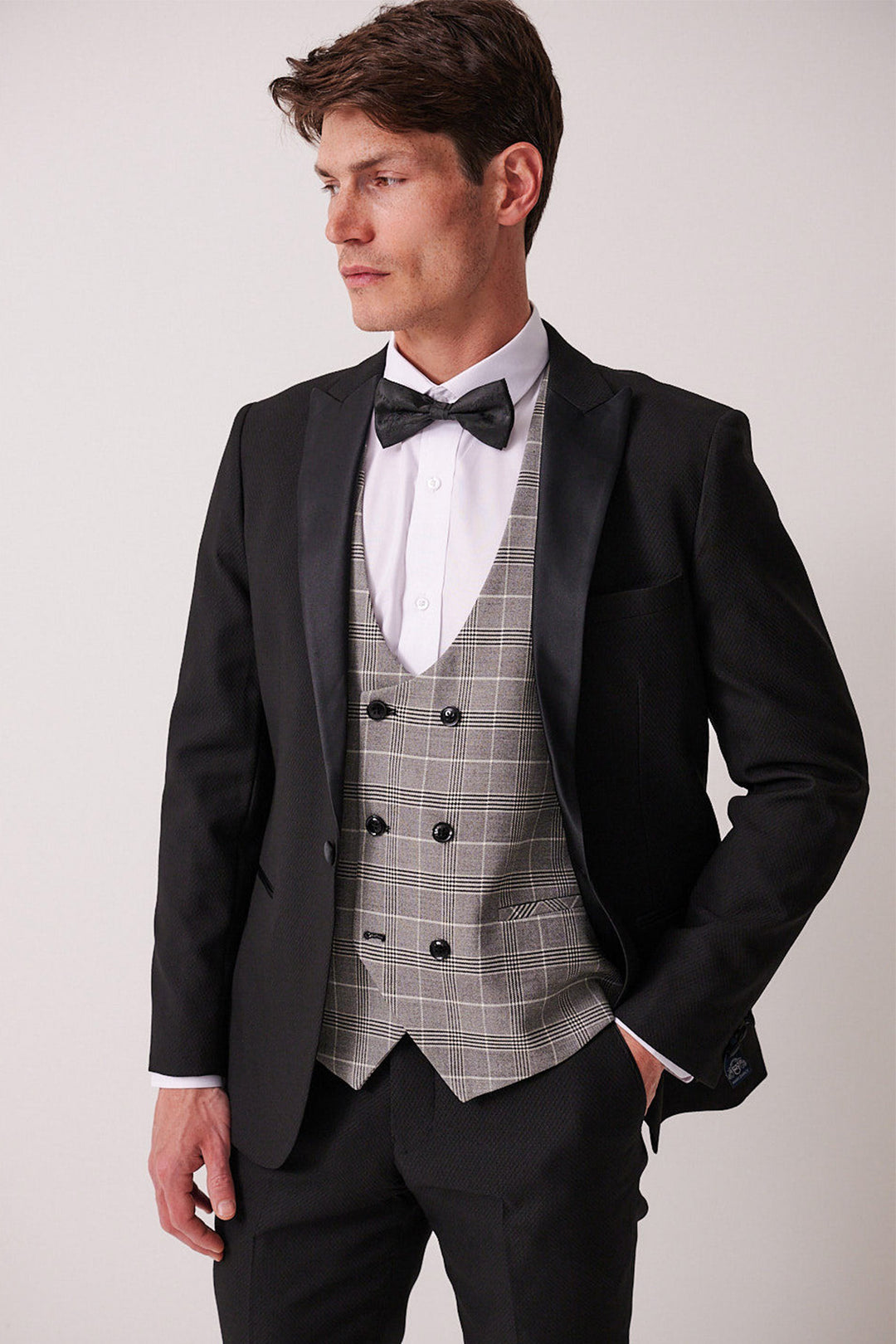 A man wearing the marcdarcy DALTON Black Tux Lapel Suit with Ross Waistcoat stands with one hand in his pocket, gazing to the side against a plain background.