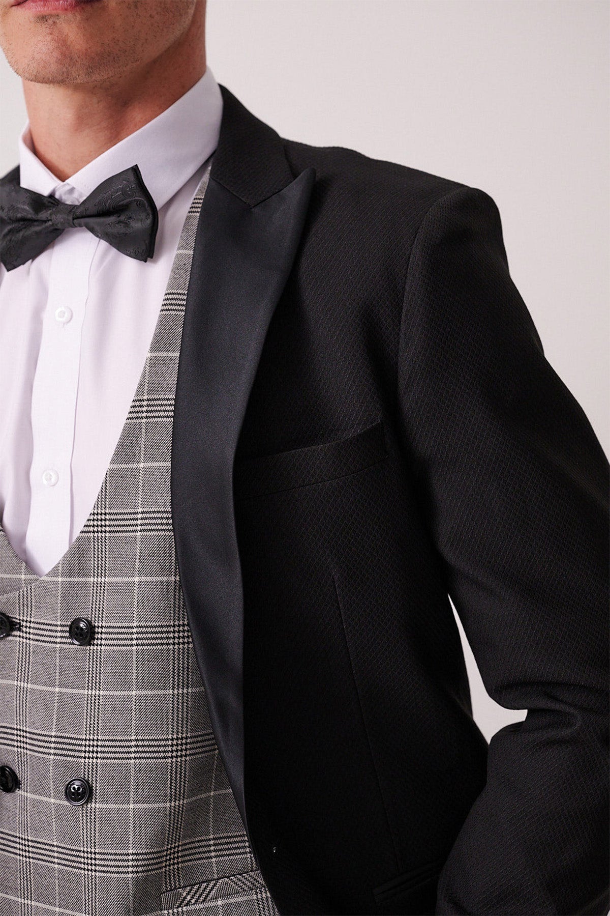 Man in the marcdarcy DALTON – Black Tux Lapel Suit with Ross Waistcoat, featuring a black patterned jacket, white shirt, black bowtie, and grey checkered waistcoat—ideal for black tie events.