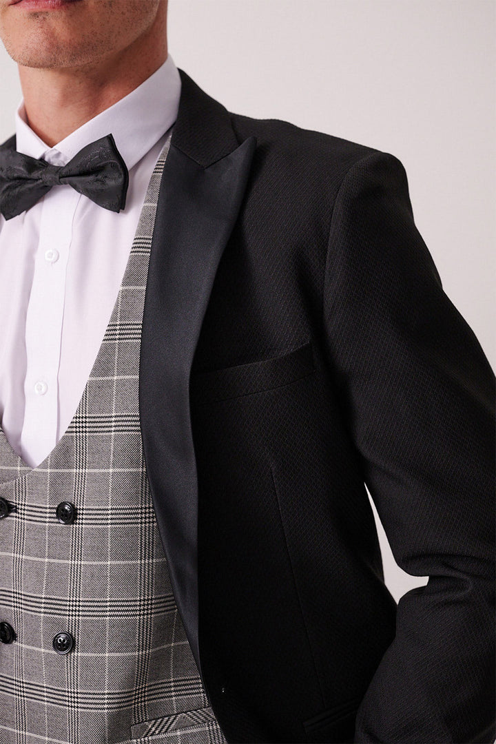 Man in the marcdarcy DALTON – Black Tux Lapel Suit with Ross Waistcoat, featuring a black patterned jacket, white shirt, black bowtie, and grey checkered waistcoat—ideal for black tie events.