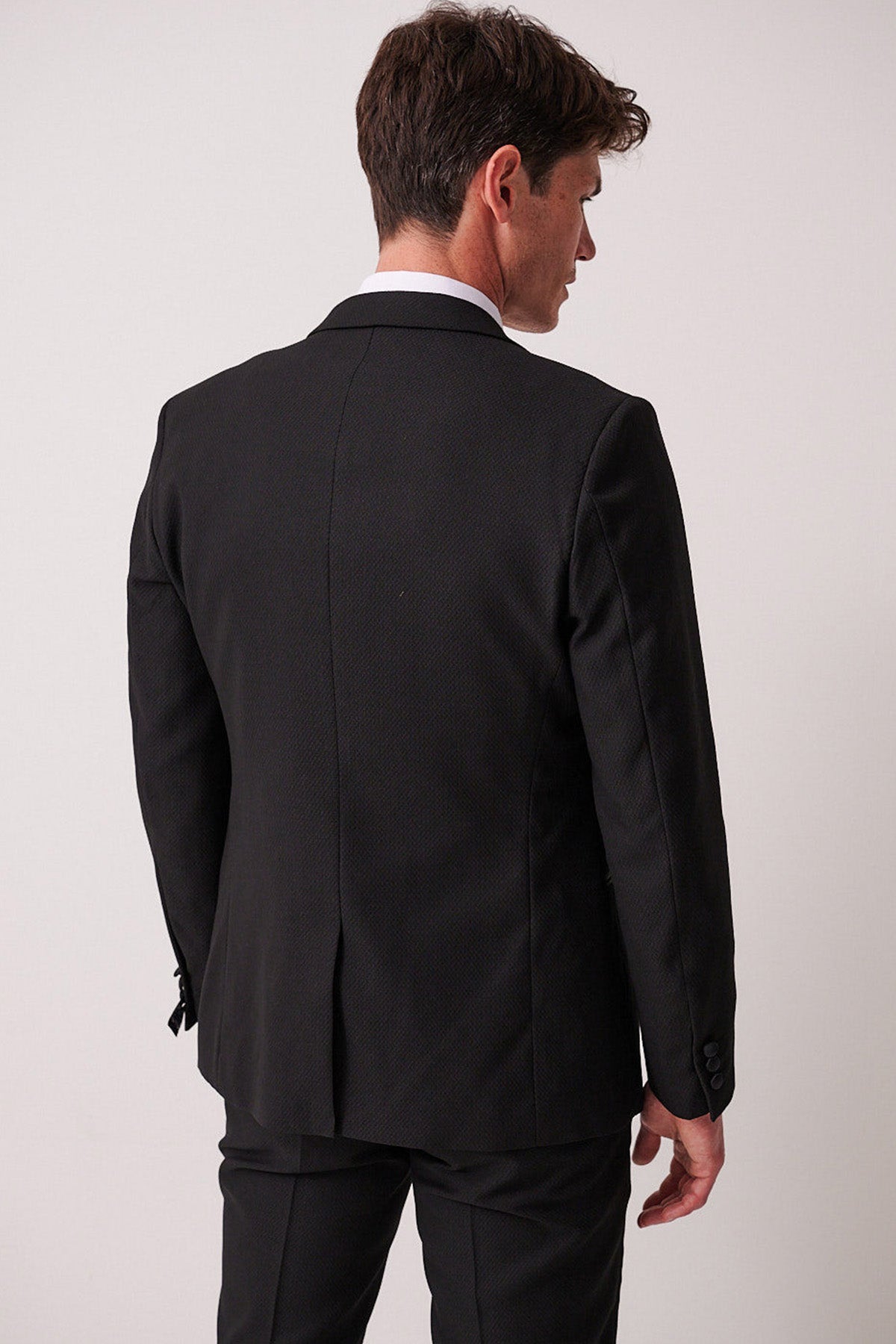 A man with short brown hair wears the marcdarcy DALTON Black Tux Lapel Suit with Ross Waistcoat, slim-fit blazer, and stands with his back to the camera against a plain light background.
