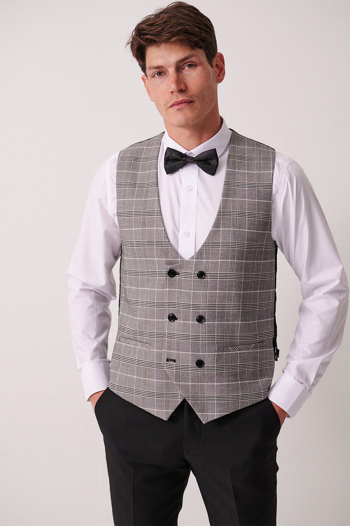 A man stands confidently in the marcdarcy DALTON Black Tux Lapel Suit with Ross Waistcoat, featuring a white dress shirt, black bow tie, and gray plaid double-breasted vest with black buttons, hands in pockets against a plain light background.