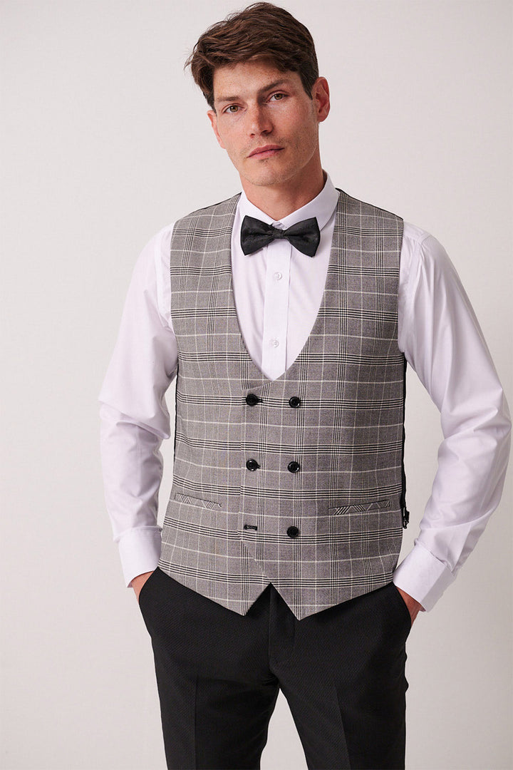 A man stands confidently in the marcdarcy DALTON Black Tux Lapel Suit with Ross Waistcoat, featuring a white dress shirt, black bow tie, and gray plaid double-breasted vest with black buttons, hands in pockets against a plain light background.