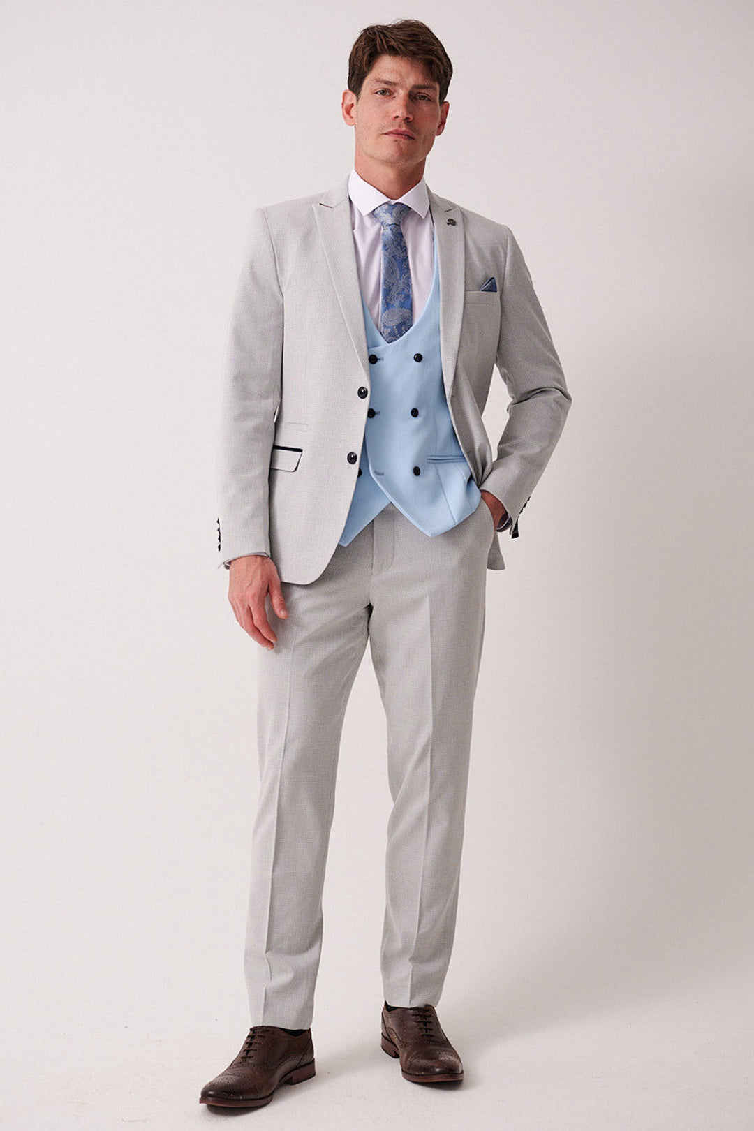 A man stands against a plain background wearing the Marc Darcy BROMLEY Stone Check Suit with Kelvin Sky Waistcoat, a white shirt, blue patterned tie, and brown shoes. His left hand is in his pocket as he looks straight ahead.