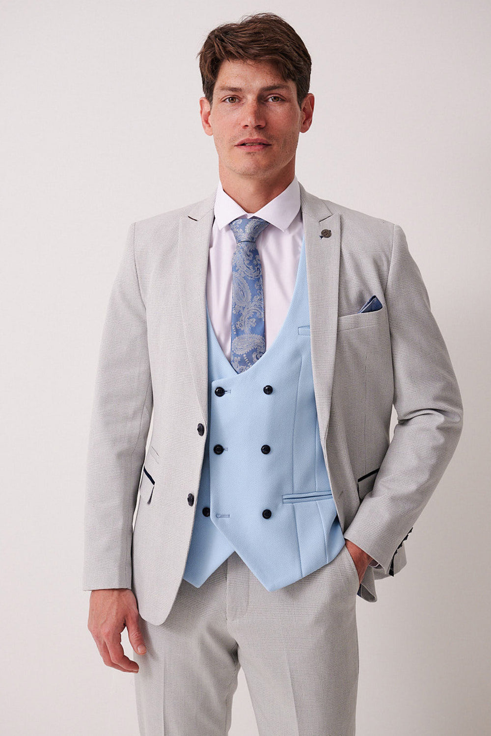 A man wears the Marc Darcy BROMLEY Stone Check Suit with Kelvin Sky Waistcoat, featuring a light gray check jacket, double-breasted light blue vest, white shirt, patterned blue tie, and pocket square against a plain backdrop.