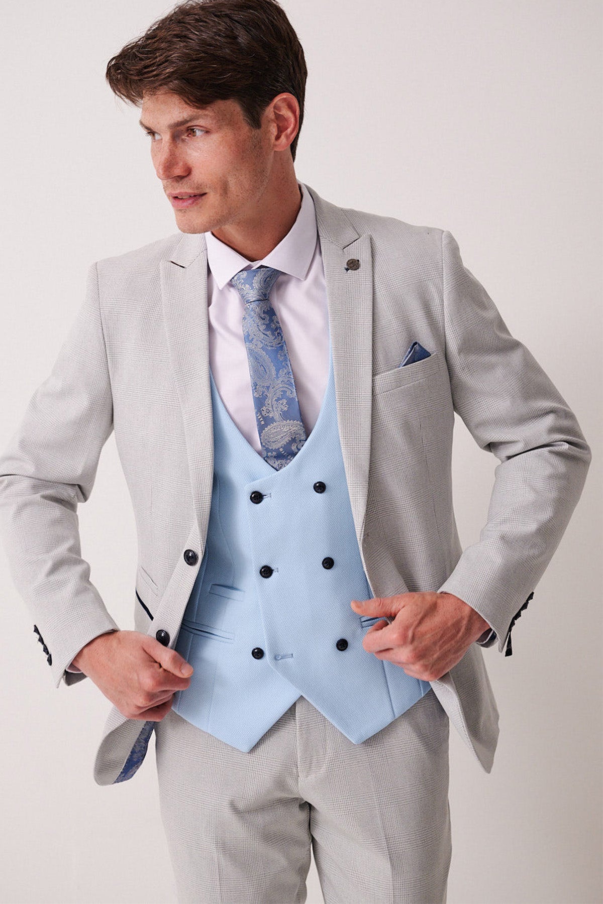 A man wears the Marc Darcy BROMLEY stone check suit with a Kelvin Sky waistcoat, paired with a white shirt and blue paisley tie.