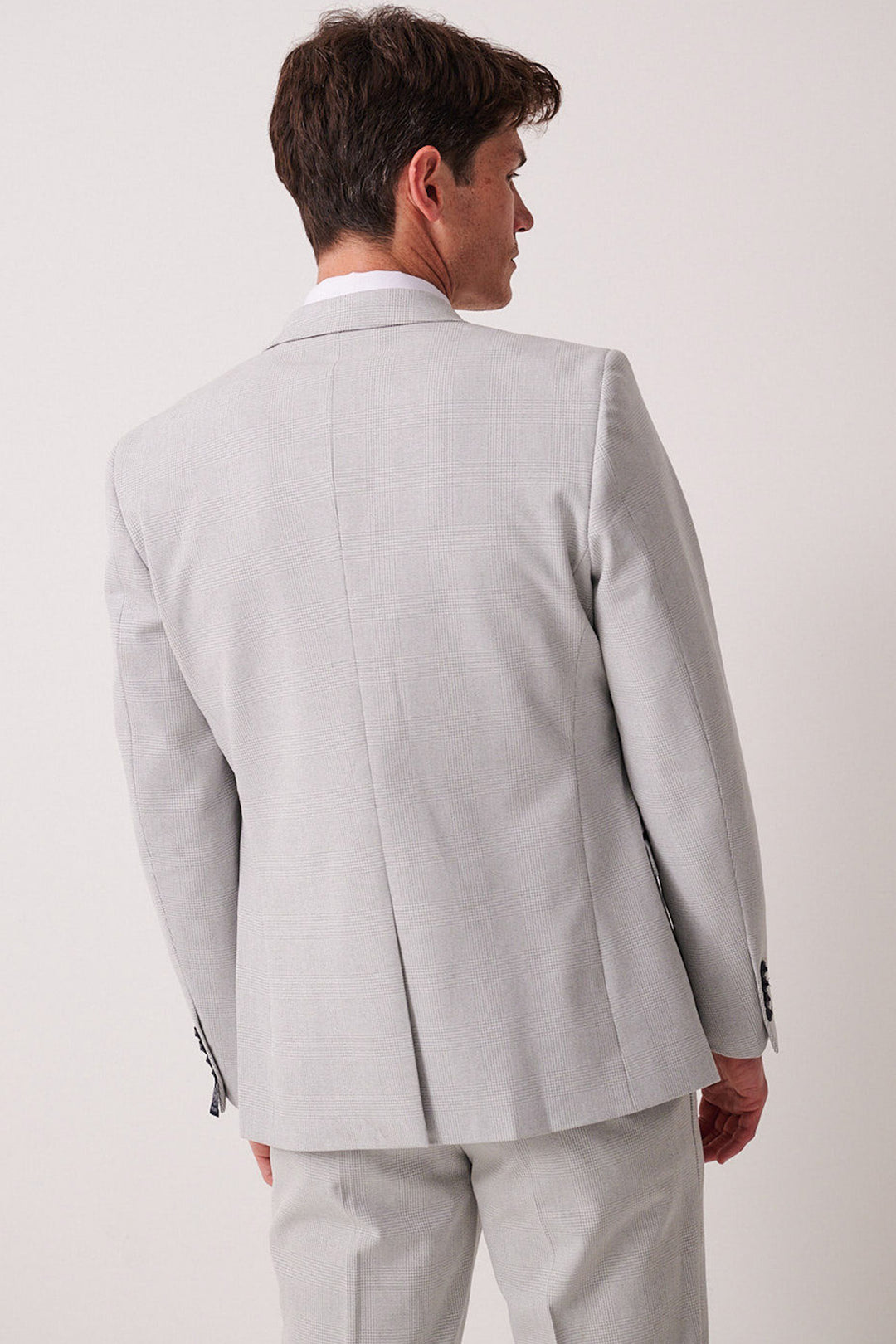 A man in a slim fit Marc Darcy BROMLEY stone check three piece suit with Kelvin Sky waistcoat stands with his back to the camera, head turned slightly to the side, against a plain light background.