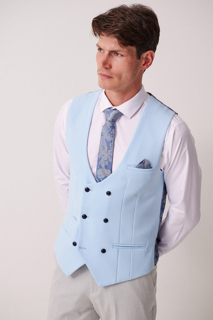 A man models the Marc Darcy BROMLEY Stone Check Suit with Kelvin Sky Waistcoat, paired with a white shirt, blue patterned tie, and light gray trousers. He stands hands behind his back, looking slightly to the side.