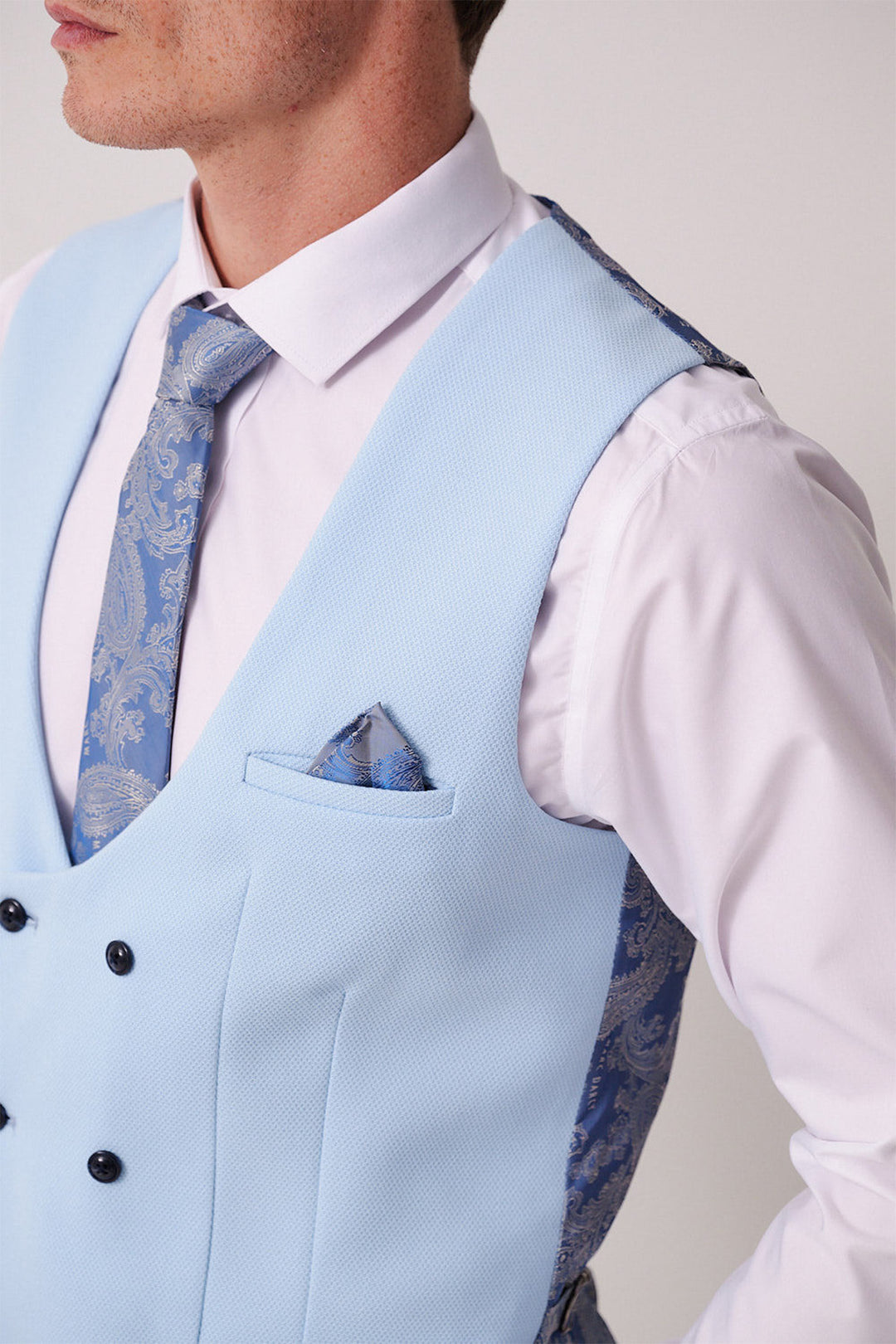 A man wears the Marc Darcy BROMLEY Stone Check Suit, paired with the Kelvin Sky Waistcoat, white shirt, and blue paisley tie and pocket square for a refined, polished three-piece look.