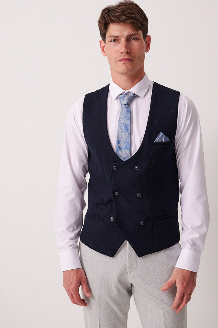 A man in Marc Darcy's BROMLEY Stone Check Suit with a Kelvin Navy Waistcoat stands against a plain light background, wearing a white shirt, blue patterned tie, and matching pocket square.