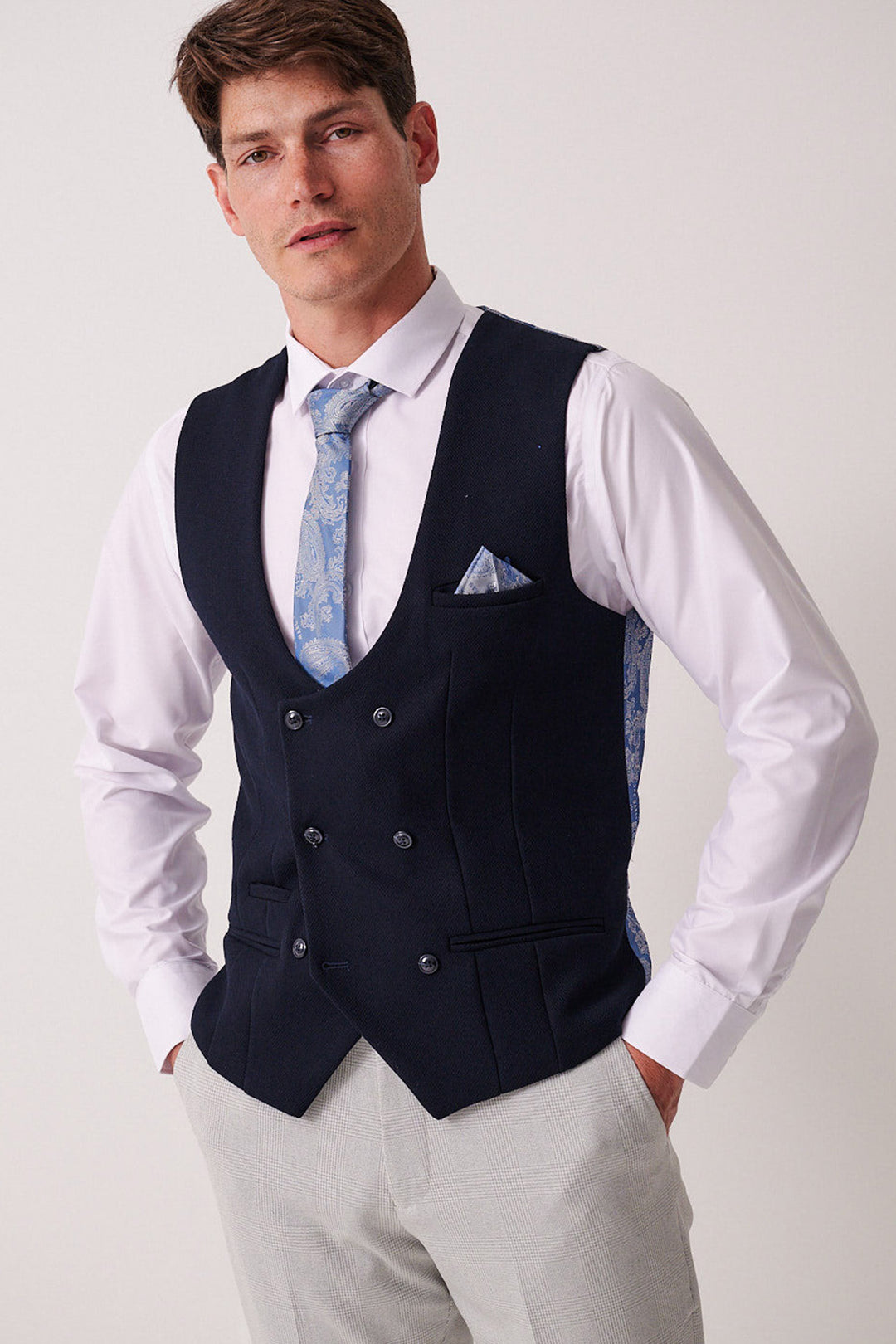 A man stands with hands in pockets, wearing the Marc Darcy BROMLEY Stone Check Suit paired with a Kelvin Navy Waistcoat over a white shirt and light blue patterned tie, finished with a matching pocket square, against a plain background.