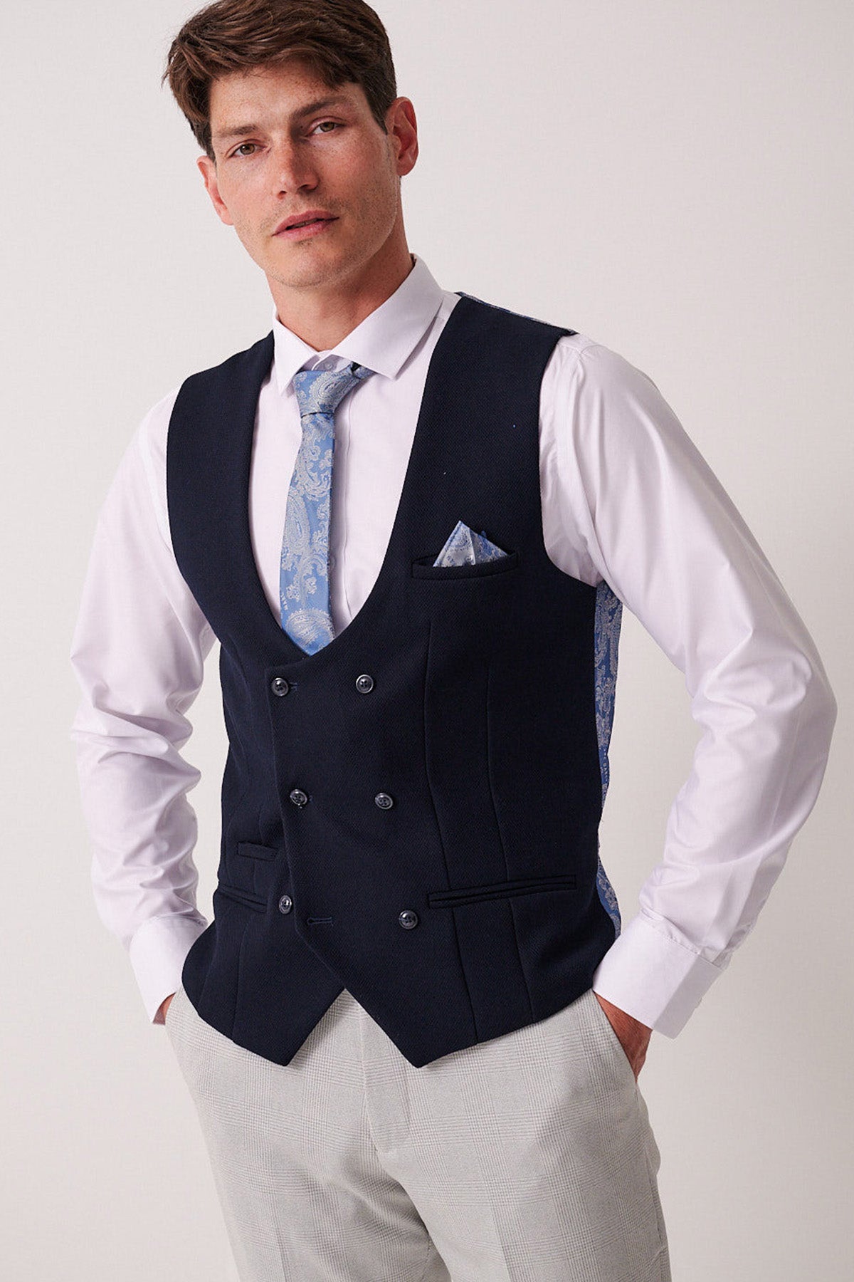 A man stands with hands in pockets, wearing the Marc Darcy BROMLEY Stone Check Suit paired with a Kelvin Navy Waistcoat over a white shirt and light blue patterned tie, finished with a matching pocket square, against a plain background.