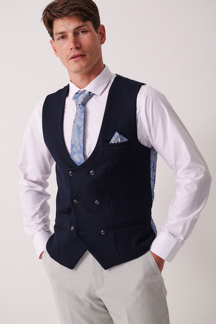 A man stands with hands in pockets, wearing the Marc Darcy BROMLEY Stone Check Suit paired with a Kelvin Navy Waistcoat over a white shirt and light blue patterned tie, finished with a matching pocket square, against a plain background.