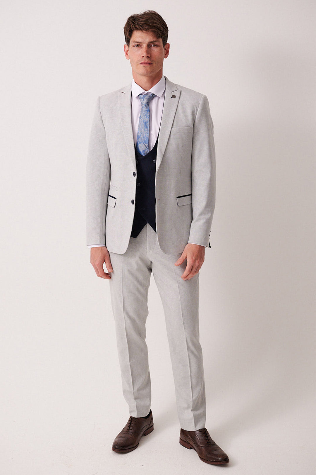 A man with short brown hair and a neutral expression wears the Marc Darcy BROMLEY Stone Check Suit with a Kelvin Navy Waistcoat, white shirt, blue patterned tie, and brown dress shoes against a plain light background.