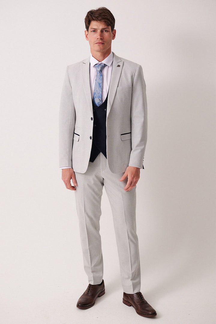 A man with short brown hair and a neutral expression wears the Marc Darcy BROMLEY Stone Check Suit with a Kelvin Navy Waistcoat, white shirt, blue patterned tie, and brown dress shoes against a plain light background.