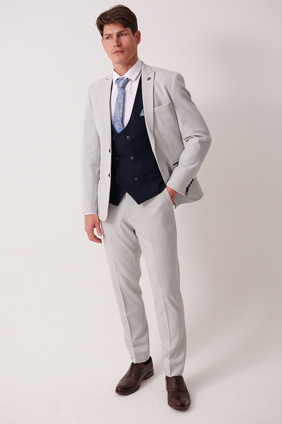 A man wears the Marc Darcy BROMLEY Stone Check Suit with Kelvin Navy Waistcoat, styled with a white shirt, light blue patterned tie, and brown dress shoes against a plain white background.