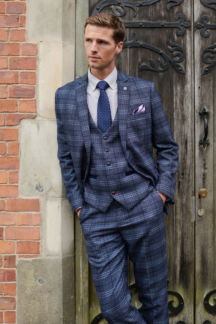 Wearing the Marc Darcy ANDY Blue Check Three Piece Suit, a man stands by a brick and stone wall near a wooden door, looking to the side with his hands in his pockets.
