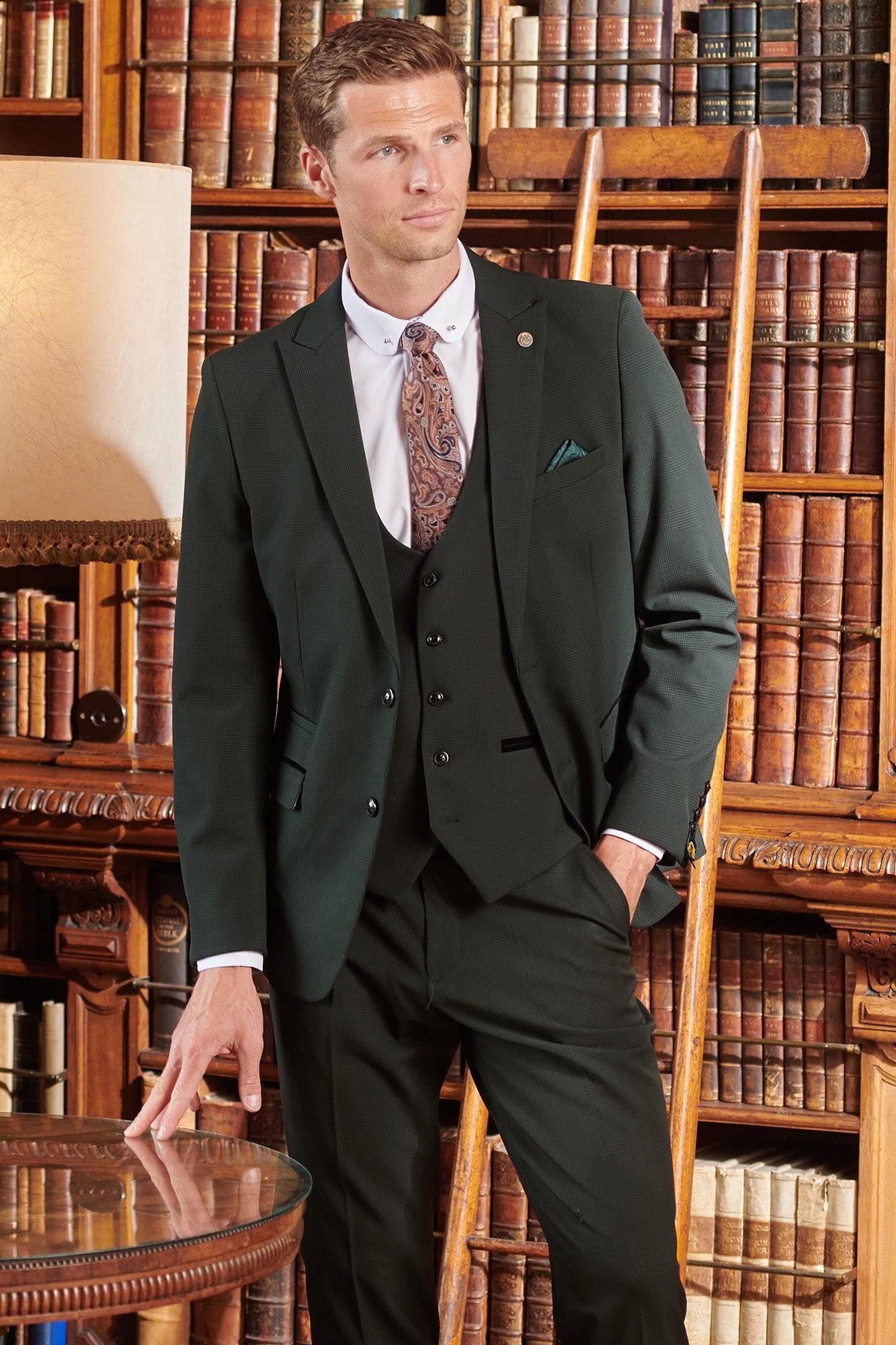 A man in a Marc Darcy BROMLEY Olive Green Check Three Piece Suit stands by bookshelves, one hand in his pocket and the other on a glass table, with a wooden ladder and lamp nearby—ideal attire for formal occasions.