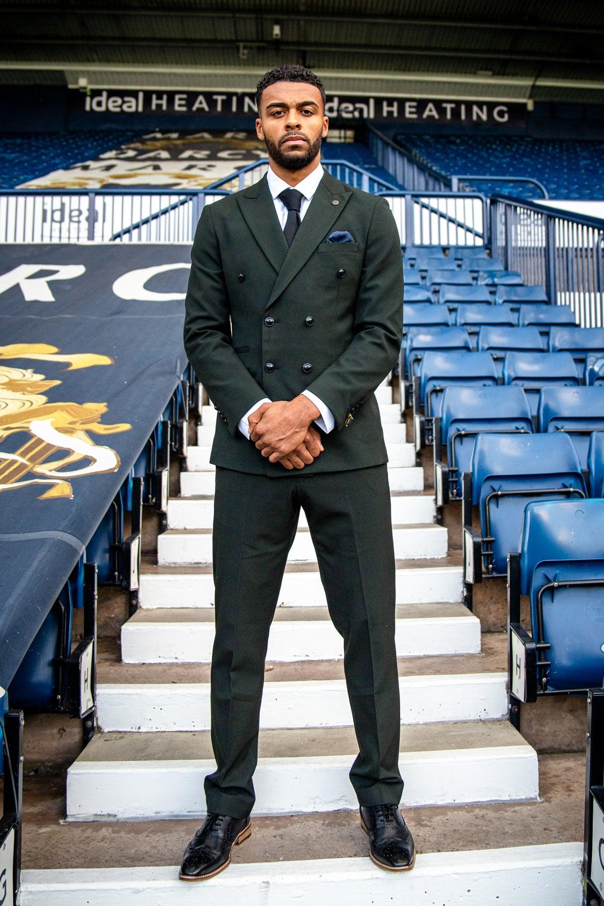 A man in The WBA Collection - BROMLEY Olive Green Double Breasted Suit by Marc Darcy, as worn by Darnell Furlong, stands on stadium stairs.